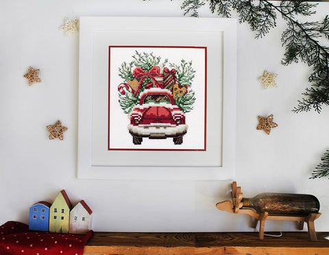 Siuvinėtas paveikslas: raudonas senovinis automobilis su Kalėdų dovanomis ir eglutės šakomis, baltame rėme 30×30 cm
