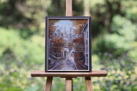 Kryželiu siuvinėtas paveikslas rėme ant molberto, rudeniškos miesto gatvės motyvas, 30×40 cm, priekio kampu rodoma kompozicija