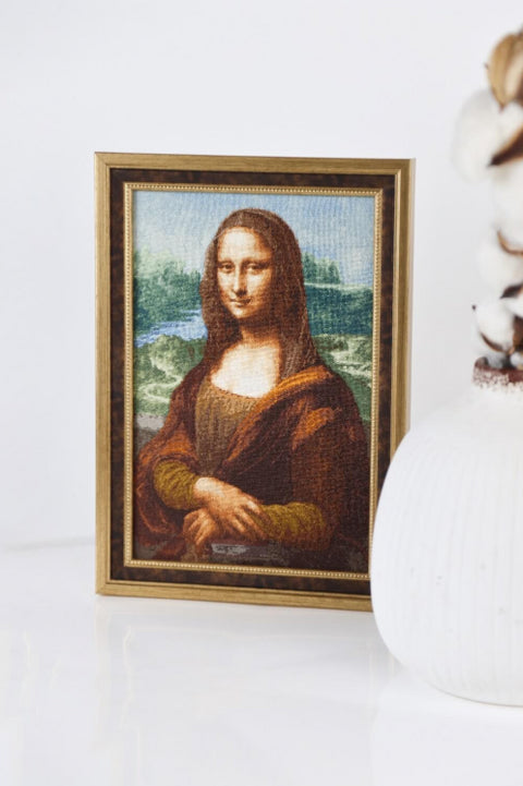 Siuvinėtas paveikslas Mona Liza su siauru auksiniu rėmu, stovintis ant stalviršio šonu matomu kampu