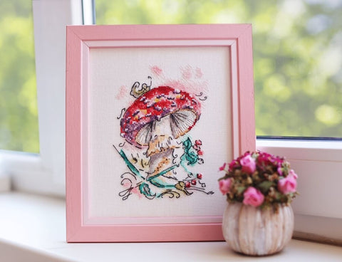 Siuvinėtas paveikslas su raudonu grybuku, rožiniu mediniu rėmu (apie 20×25 cm), ant palangės šonu fotografuotas kartu su mažyčiu rožių vazonėliu