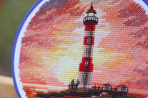 Kryželiu siuvinėtas švyturys, artimas kampas su oranžiniu ir geltonu saulėlydžio fonu, matoma iškilusi tekstūra ir smulkios siūlės