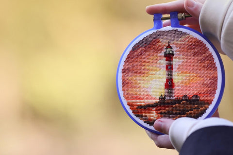 Kryželių siuvinys su raudono ir oranžinio atspalvių švyturio motyvu, rankose įtemptas mėlyname plastikiniame lankelyje, artimas planas