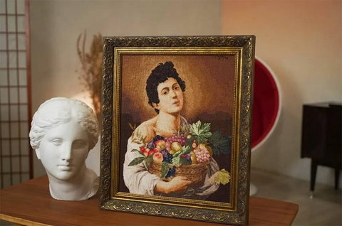 Siuvinėtas paveikslas kryželiu — klasikinis jauno žmogaus su vaisių krepšeliu motyvas auksiniame ornamentuotame rėme, 3/4 kampu ant stalo