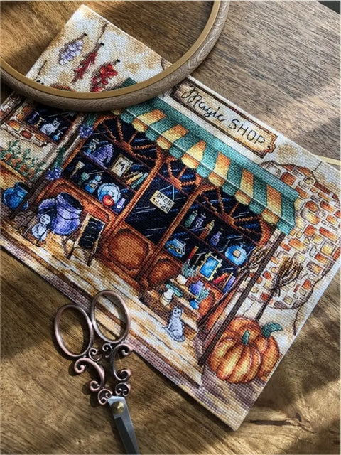 Kryželių siuvinėjimo rinkinys – rankų darbo „Magic Shop“ motyvas arti kadro, matomas su siuvimo lankeliu ir dekoratyvinėmis žirklėmis