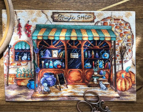 Siuvinėta drobė "Magic Shop" mažo formato, frontali kompozicija su žaliu tentu, rudeniškais moliūgais ir priekyje žirklių rekvizitu