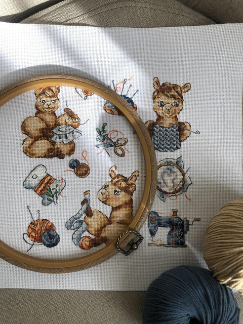 Siuvinėjimo rinkinys su alpakių motyvais, parodytas arti su dekoratyviniu mediniu lankeliu ir pilkais bei smėlio spalvos siūlais