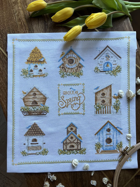 Kryželiu siuvinys Hello Spring – kvadratinis apie 30×30 cm baltame audinyje su devyniais geltonais ir mėlynais paukščių nameliais, fotografuotas iš viršaus