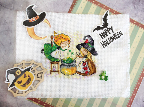 Kryželių siuvinys: Halloween motyvas — dvi raganiukės prie katilo, žalia‑oranžinė spalvų schema ant balto Aida, fotografuota iš viršaus