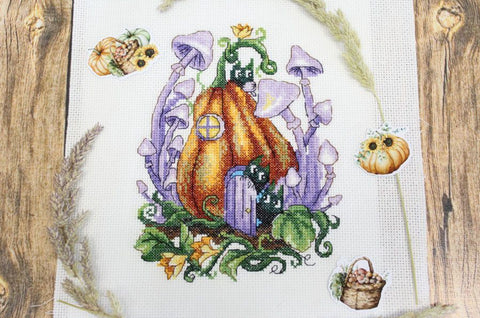 Siuvinėjimo rinkinys su kryželiu: moliūgo namelis oranžinėmis ir violetinėmis detalėmis ant balto audeklo, pateiktas flatlay su kaimiškomis dekoracijomis