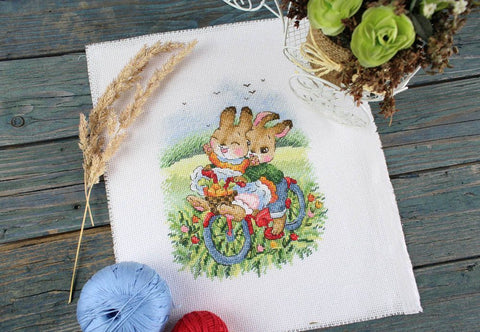 Kryželių siuvinys „Zuikiai ant dviračio“ ant balto audinio ~20×20 cm, plokščias produktų kadravimas su dekoratyviais siūlais ir gėlėmis