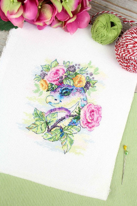 Kryželių siuvinėjimo rinkinys: pastelinės spalvos dinozauras su gėlių vainiku ant balto audinio, plokštuminis artimas kadras, 20×25 cm