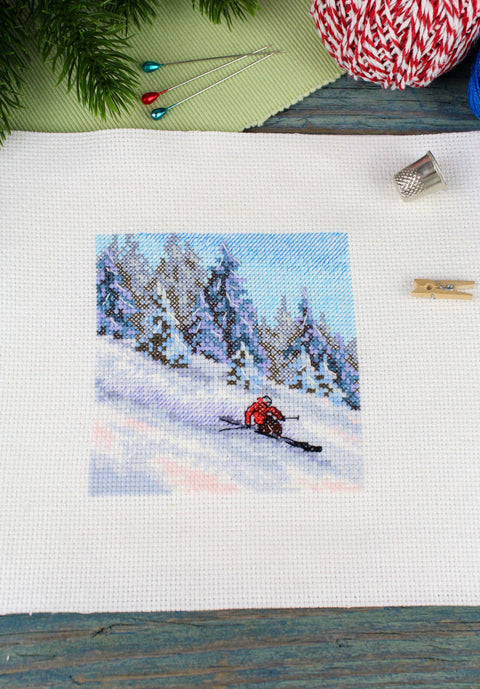 Siuvinėjimo rinkinys su žiemos slidininko motyvu ant balto Aida audinio (dydis ~10x10 cm), parodytas iš viršaus su raudinu slidininko siuviniu ir siuvimo įrankiais