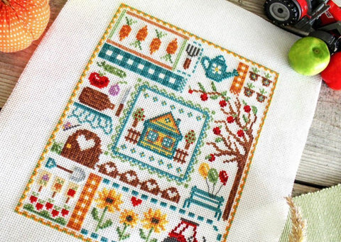 Kryželių siuvinys Country Sampler ant balto audinio su nameliu centre, obelimi, saulėgrąžomis ir daržo motyvais