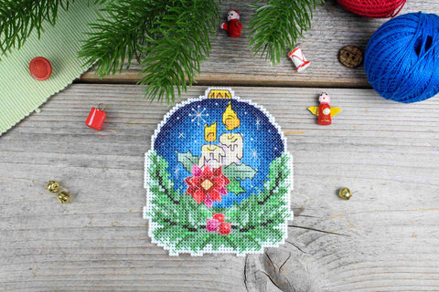 Siuvinėtas kalėdinis ornamentas su dviem žvakėmis, tamsiai mėlynu fonu ir raudona žvaigždute; 10×8 cm, priekio rodinys ant medinės lentos, rankų darbo detalė