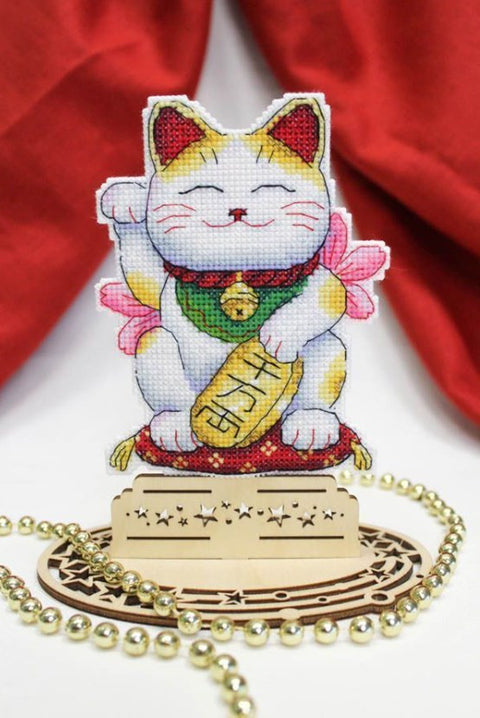 Maneki-neko siuvinėta statulėlė, balta su auksinėmis dėmėmis ir žaliu kaklaraiščiu, ant medinio žvaigždžių pagrindo