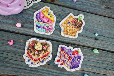 Siuvinėta sagė rinkinys: keturios spalvotos desertų formos (cupcake, vaflis, tortas, pyragas), rankų darbo su karoliukų detalėmis, 4–5 cm, padėtos ant medinio fono, unikalios kiekvienos