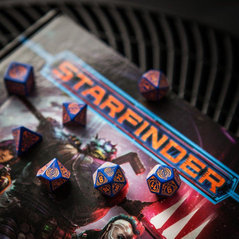 RPG kauliukai mėlyni su vario skaičiais komplekte, fotografuoti ant Starfinder žaidimo taisyklių knygos viršelio