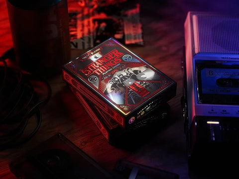 Stranger Things kortų rinkinys su raudro‑juodo blizgaus dėklo dizainu, trijų dėžių stulpelyje šone, fone senovinė kasetinė grotuvas
