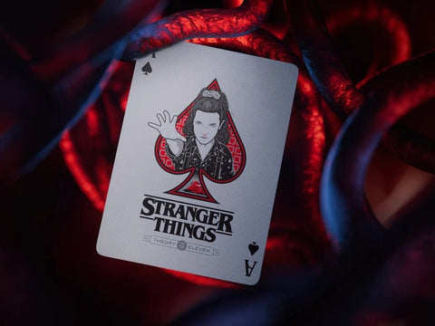 Stranger Things žaidimo korta tūzas su Eleven iliustracija, raudono‑juodos detalės, pasvirusiu kampu prieš tamsias raudonas vijokles