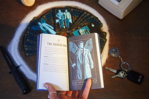 Tarot knyga atversta prie The Hanged Man paaiškinimo, rankoje laikomas kišeninis iliustruotas vadovas, fone išskleistos kortos