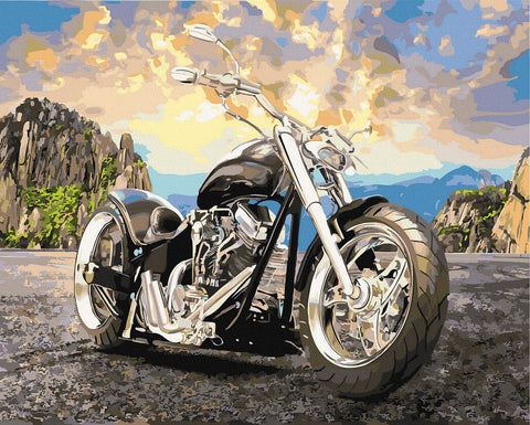 Motociklas chopper juodas, priekiniu kampu parodytas su chromuotu varikliu ir dideliu ratu, fone kalnų saulėlydis