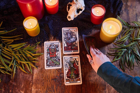 Tarot kortos rinkinys ant medinio stalo su degančiomis žvakėmis, kaukolės dekoracija ir ranka atverčia kortą