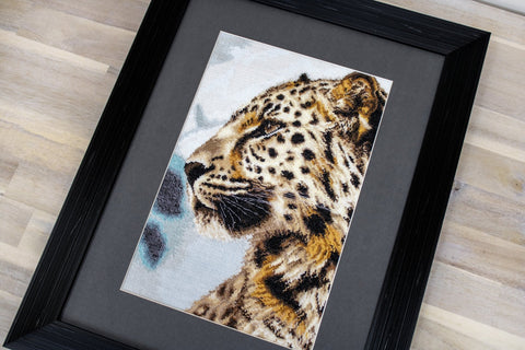 Įrėmintas siuvinys leopardo profiliu su juodu mediniu rėmu, detalus kampinis artinimas rodo drobės tekstūrą ir dėmių raštą