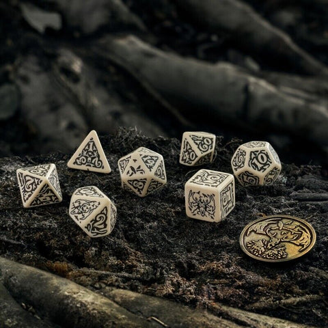 RPG kauliukai: 7 dalių dramblio kaulo spalvos rinkinys su juodais ornamentais, išdėstyti ant medžio žievės su metaline moneta masteliui