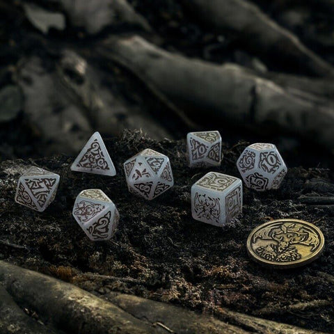 Kauliukai RPG rinkinys: balti su tamsiomis ornamentuotomis įgraviruotomis kraštinėmis, pateikti ant samanotos miško žemės šalia metalinės monetos