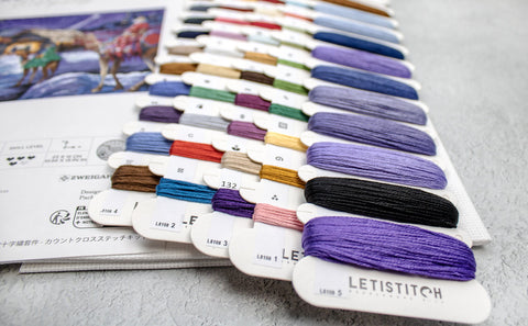 Siuvinėjimo siūlai Letistitch įvairių violetinių ir kitų atspalvių ant kortelių, artimas kampas šalia siūlomo rašto