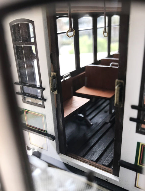 Miniatiūrinis tramvajaus vagonėlis, baltas korpusas ir tamsios grindys, arti matoma atvira durų interjero detalė
