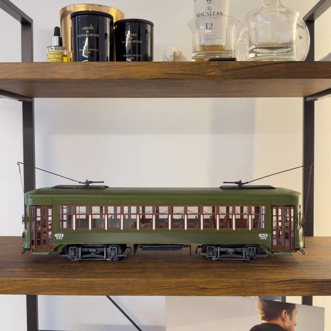 Modelinis tramvajus žaliais korpusu ir ruda langų apdaila, šoninis profilio kadravimas ant medinės lentynos, matomas sėdimų vietų interjeras