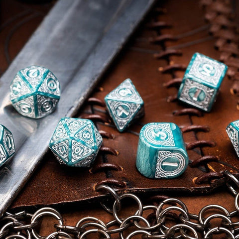 RPG kauliukai tirkiai‑sidabriniai, graviruotų 7 dalių rinkinys, artimas kadras ant odinio žaidimo kilimėlio su metaline grandinėle