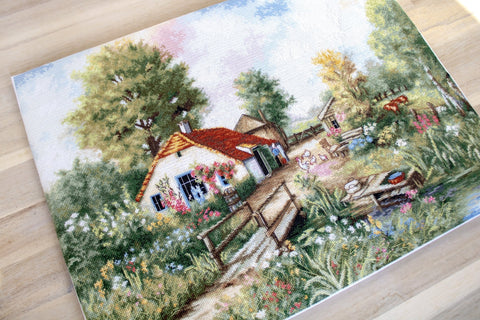 Siuvinėjimo drobė su kaimo sodybos motyvu, ryškūs žali ir raudoni tonai, 30×40 cm, artimas kampas, matomas medinis tiltelis