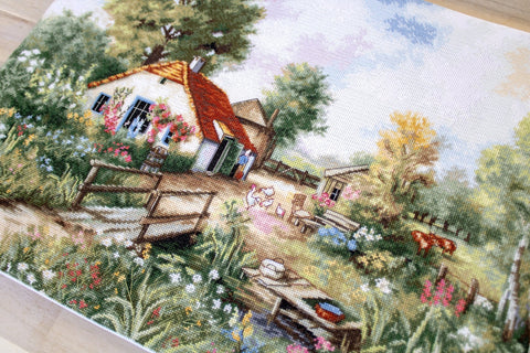 Siuvinėta drobė kaimo sodybos scena, arti kampu, ryškios spalvos ir matoma audinio tekstūra, 40×30 cm