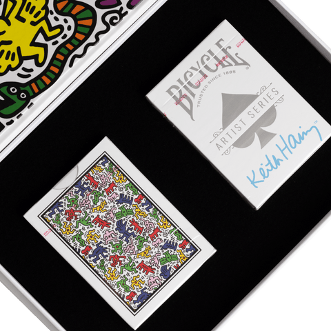 Bicycle kortos Keith Haring dovanų komplekte — baltas firmos dėklas su mėlynu parašu ir spalvotu nugarėlių raštu juodame putplasčio įdėkle