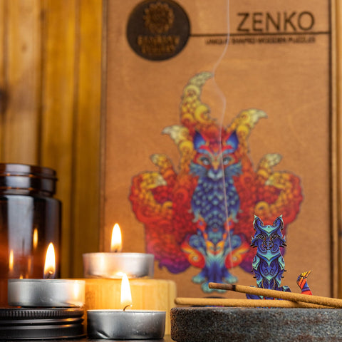 Medinė dėlionė Zenko su mėlyna–violetine lapių figūrėle prie dėžutės, uždegtos žvakės ir kvepiančių smilkalų dūmai