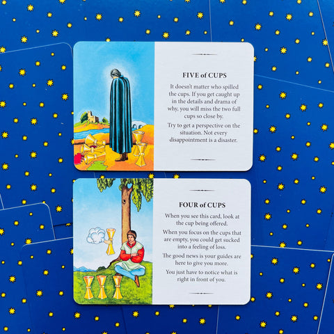 tarot kortos dvi atviros kortos (Five ir Four of Cups) su ryškiomis iliustracijomis ir žvaigždėtomis mėlynomis nugarėlėmis
