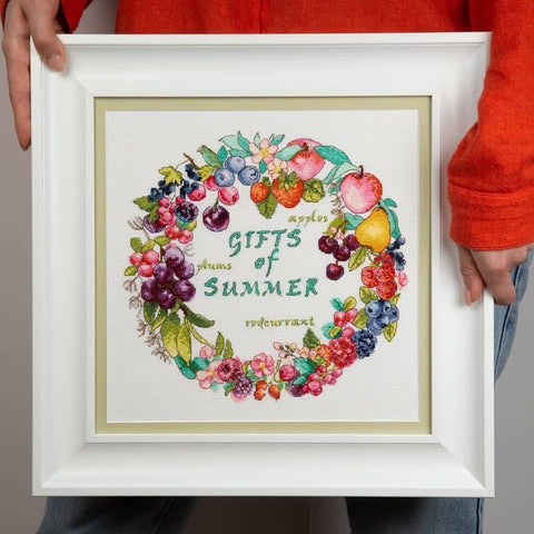 Rankų darbo siuvinėtas vaisių vainikas „Gifts of Summer“ baltame rėme, frontinis vaizdas su rankomis ir dalimi megztinio