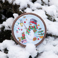 Rankų darbo kryželiu siuvinėtas žieminis sniego žmogaus ornamentas apvaliam mediniam lankeliui, raudona‑žalia šaliko detalė, fotografuota ant sniego, artimas planas