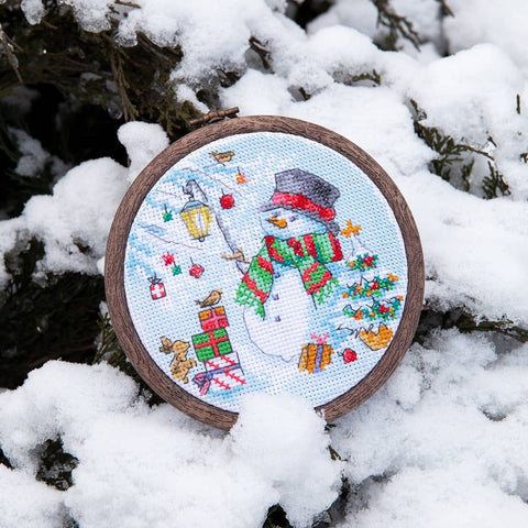 Rankų darbo kryželiu siuvinėtas žieminis sniego žmogaus ornamentas apvaliam mediniam lankeliui, raudona‑žalia šaliko detalė, fotografuota ant sniego, artimas planas