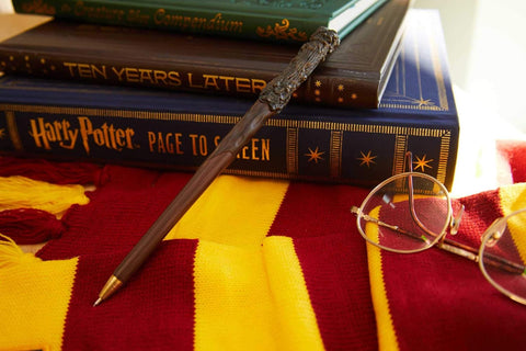 Rašiklis lazdos formos su raižytu kotu ir žalvariniu galiuku, padėtas ant raudonai–auksinio Gryffindor šaliko prie knygų