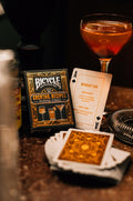 Bicycle kortų kaladė juoda auksiniu dizainu su Cocktail Recipes dėkle ir atversta Manhattan recepto kortele ant baro