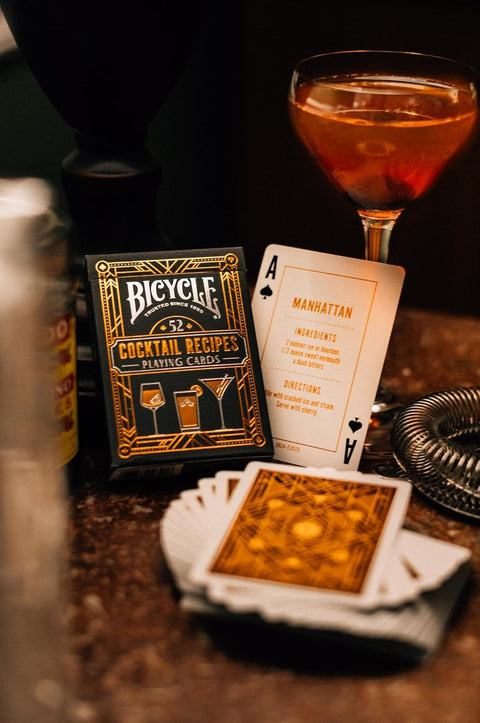 Bicycle kortų kaladė juoda auksiniu dizainu su Cocktail Recipes dėkle ir atversta Manhattan recepto kortele ant baro