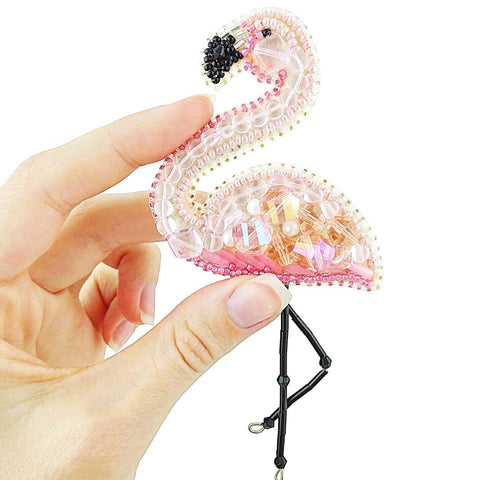 Produkto puslapyje: rožinė karoliukais ir kristalais puošta flamingo segė, artimas vaizdas, ranka laiko ir parodo dydį bei detales