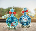 Parduodami du mėlyni siuvinėti kalėdiniai ornamentai ant medinių stovų; kairėje – snieguotas namelis, dešinėje – užrašas Enjoy