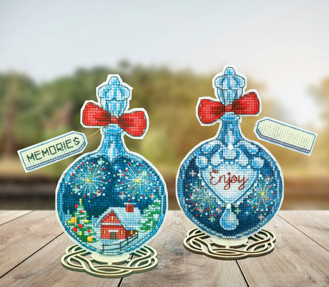 Parduodami du mėlyni siuvinėti kalėdiniai ornamentai ant medinių stovų; kairėje – snieguotas namelis, dešinėje – užrašas Enjoy