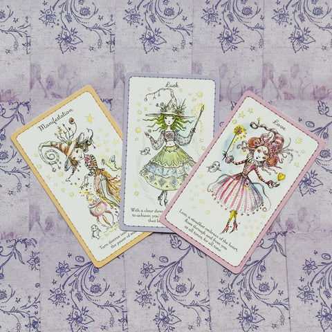 Orakulo kortų rinkinys su trimis pastelinių rėmelių kortomis (Manifestation, Luck, Love), išdėstytas įstrižai ant violetinio raštuoto fono