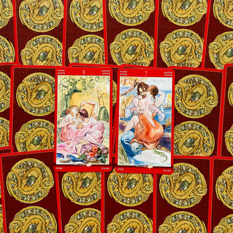 Tarot kortos raudonomis nugarėlėmis, dvi atviros kortos (7 ir 8 lazdų) su romantiškomis iliustracijomis, centruotos ant raštuoto fono