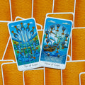 Tarot kortos Ten of Cups ir Nine of Cups su ryškiomis mėlynomis iliustracijomis ant oranžinių nugarėlių, parodytos galerijos priekinėje pozicijoje